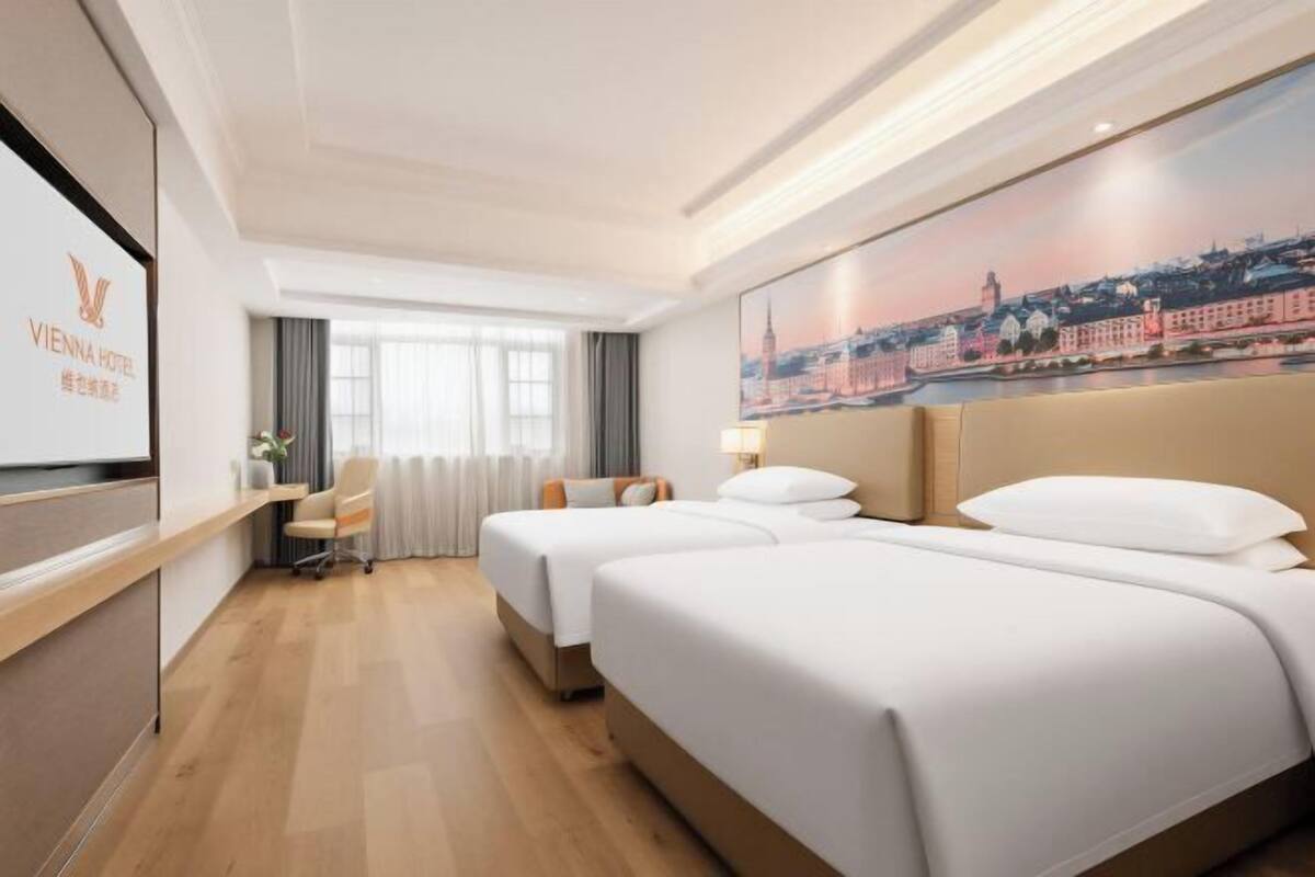 A hotel room with two beds, a desk, a chair, a TV, and a painting of a cityscape.