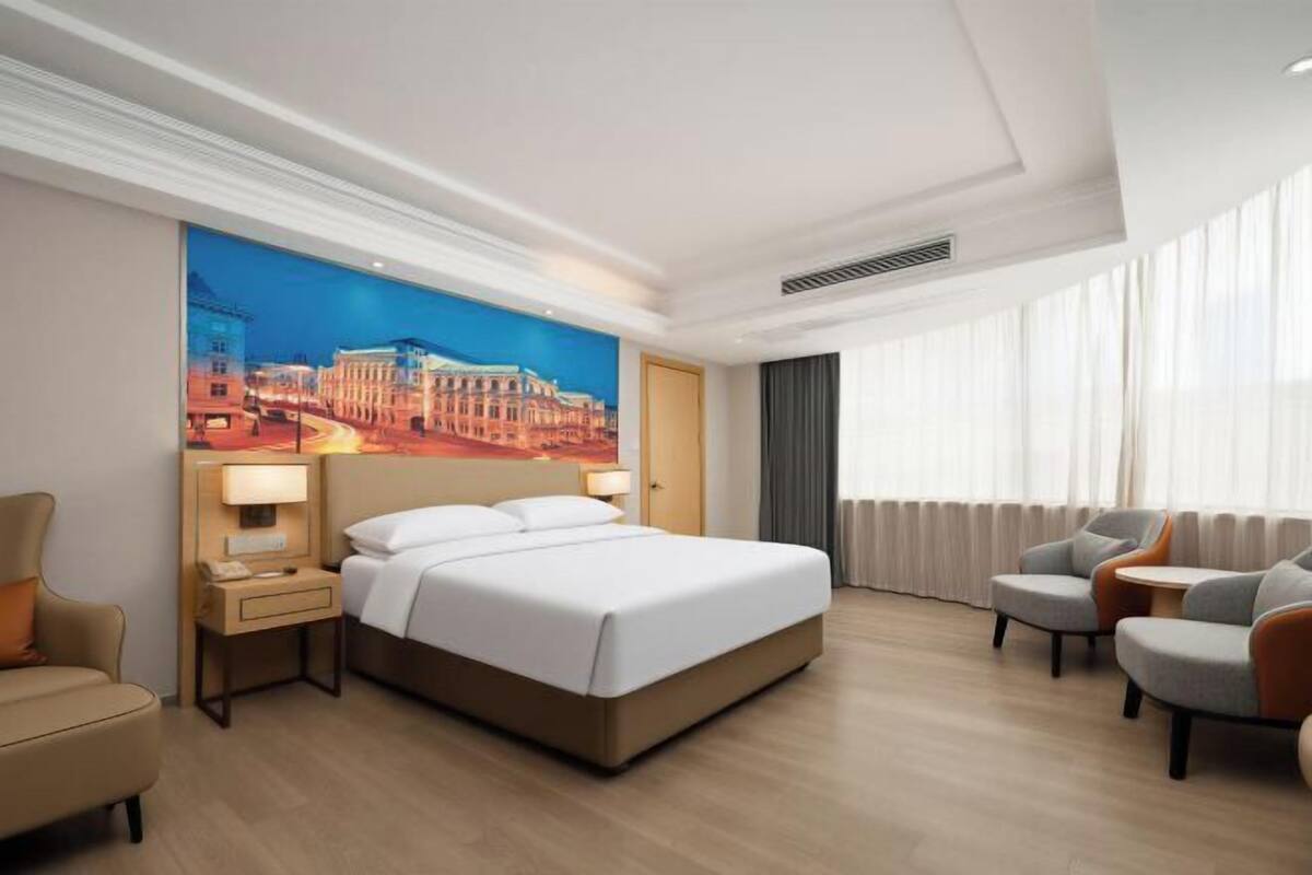 A hotel room with a bed, a nightstand, a lamp, a chair, and a view of a cityscape through the window.
