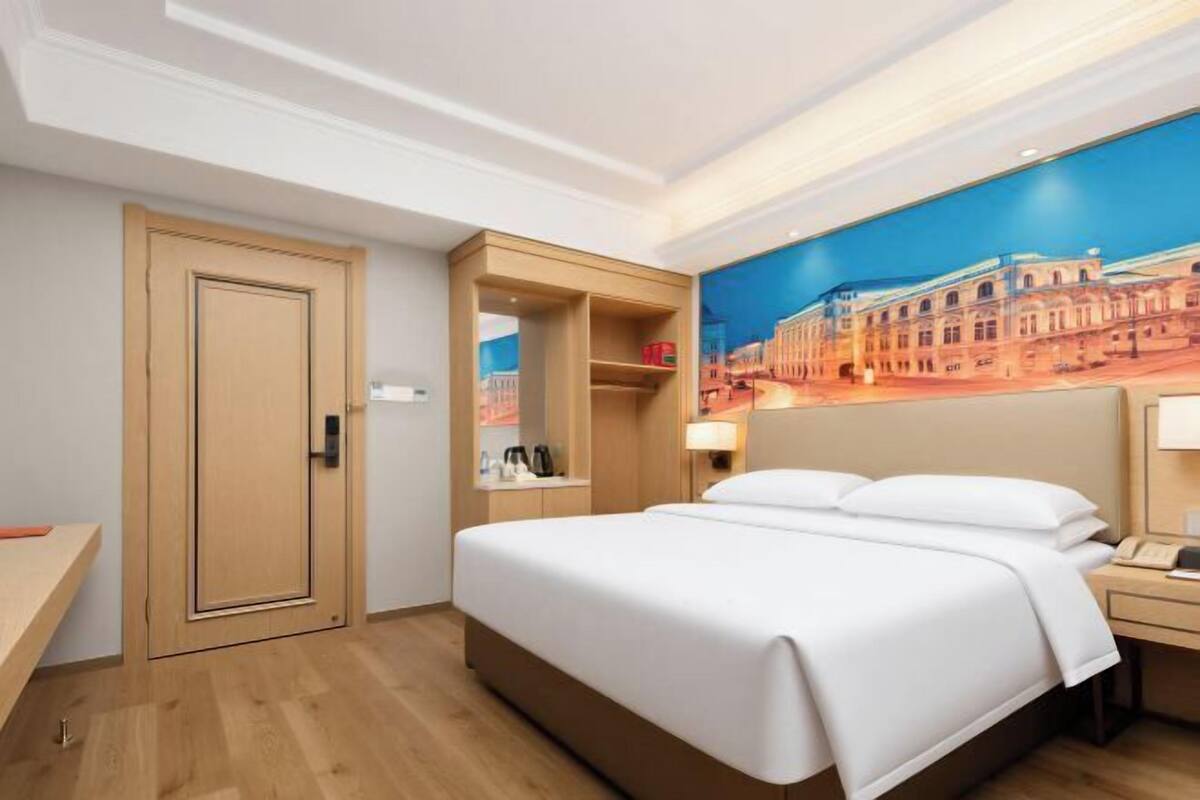 A hotel room with a bed, a desk, a wardrobe, and a painting of a cityscape on the wall.