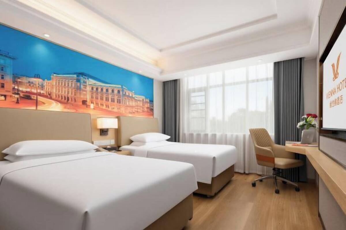 A hotel room with two beds, a desk, a chair, a lamp, and a large window.