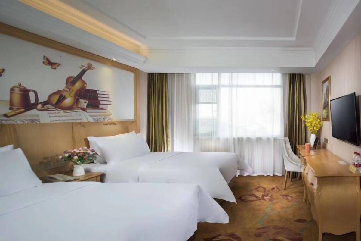 A hotel room with two beds, a desk, a TV, and a large window with curtains.
