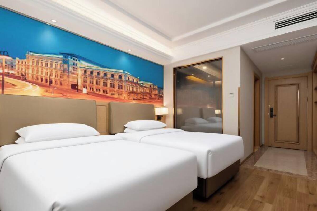 A hotel room with two beds, a large mural of a building, a wooden floor, and a door leading to another room.