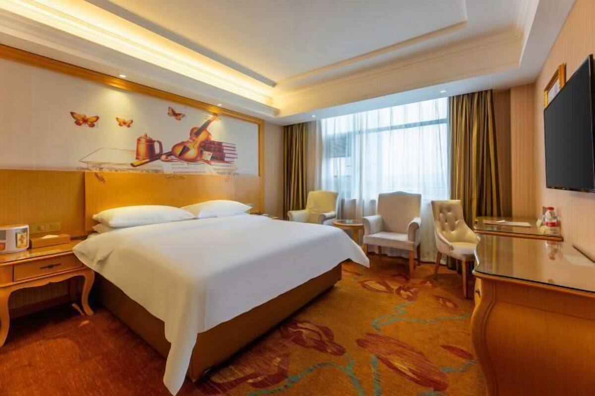 A hotel room with a large bed, a desk, a chair, a TV, and a window with curtains.