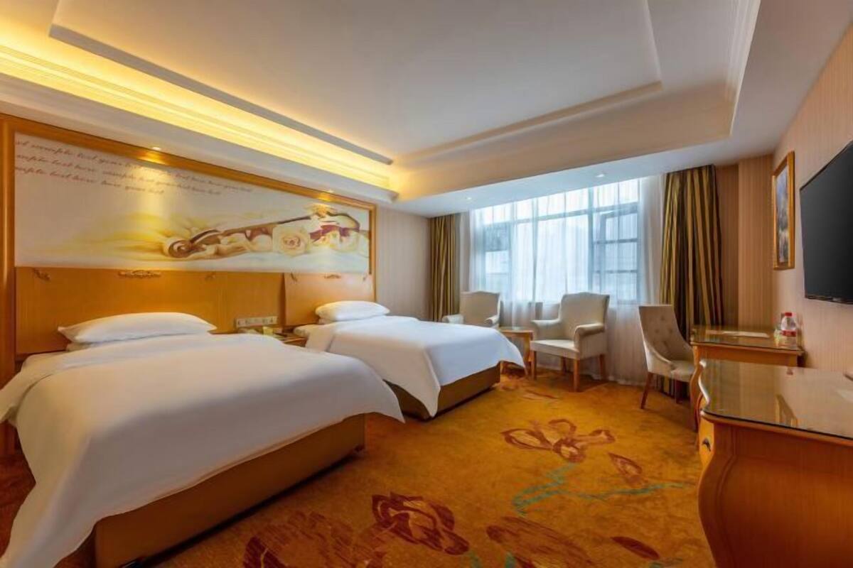 A hotel room with two beds, a desk, a chair, a TV, and a large window with curtains.