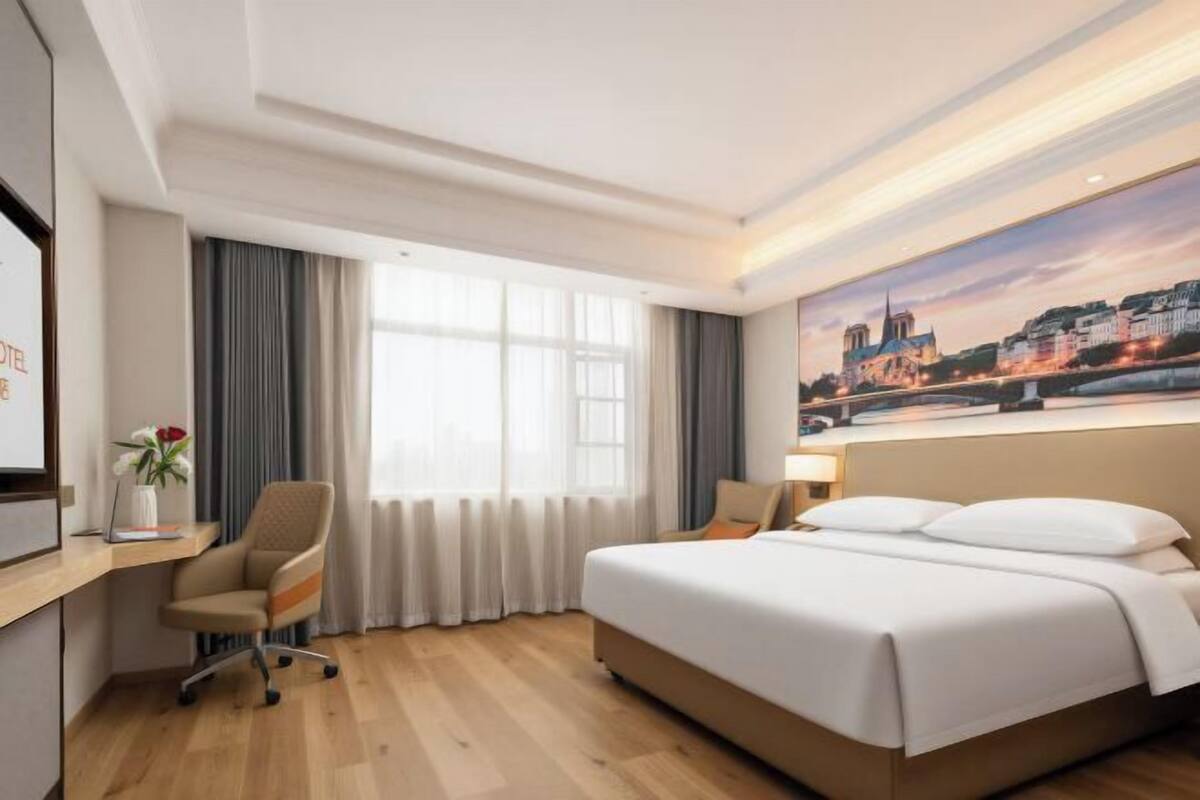 A hotel room with a large bed, a desk with a chair, a window with curtains, and a painting of a cityscape.