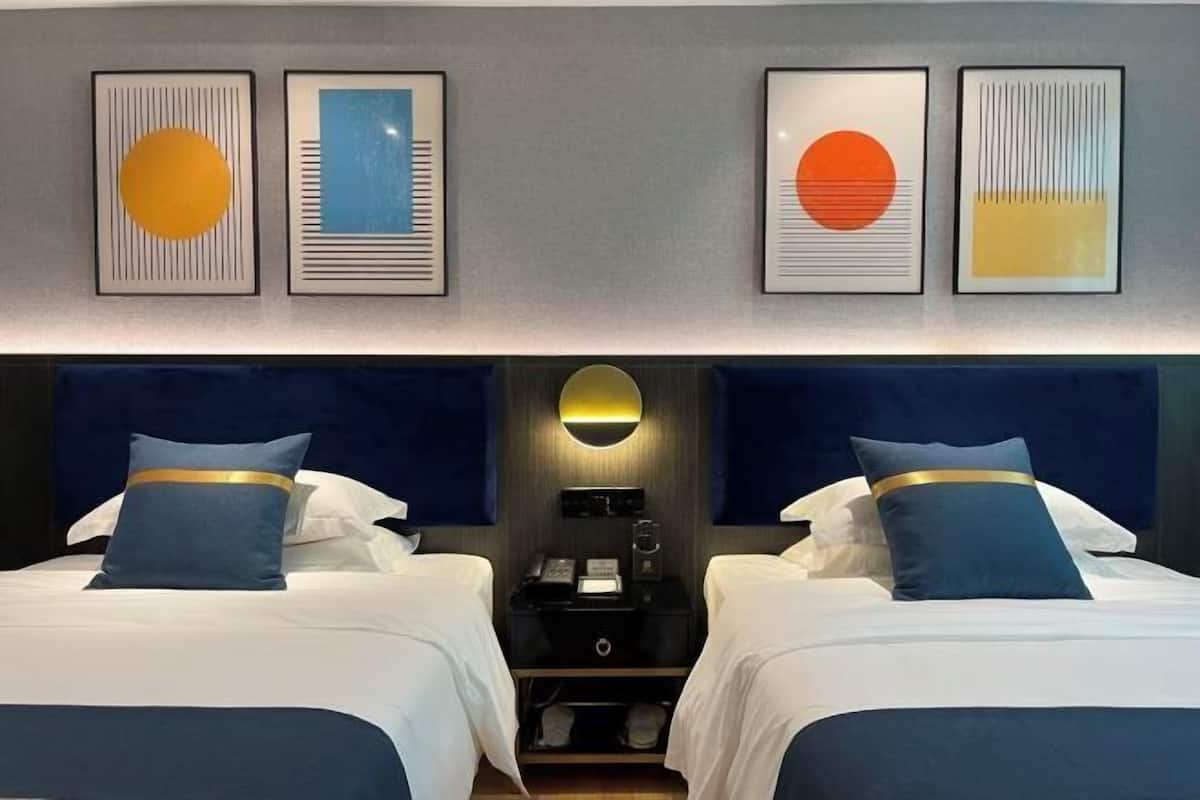Habitación de hotel con dos camas, una mesita de noche, una lámpara de pared y cuadros enmarcados sobre las camas.