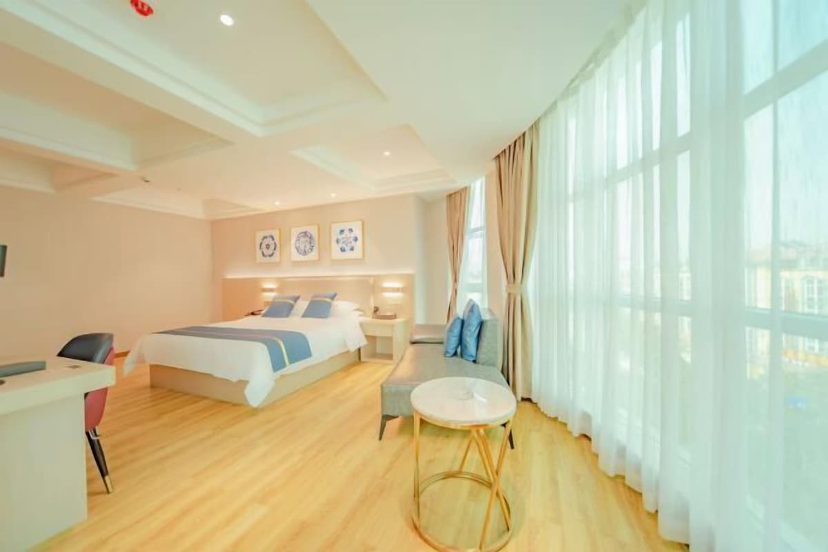 Quarto de hotel com cama, escrivaninha, cadeira, mesa pequena e janelas amplas com cortinas.