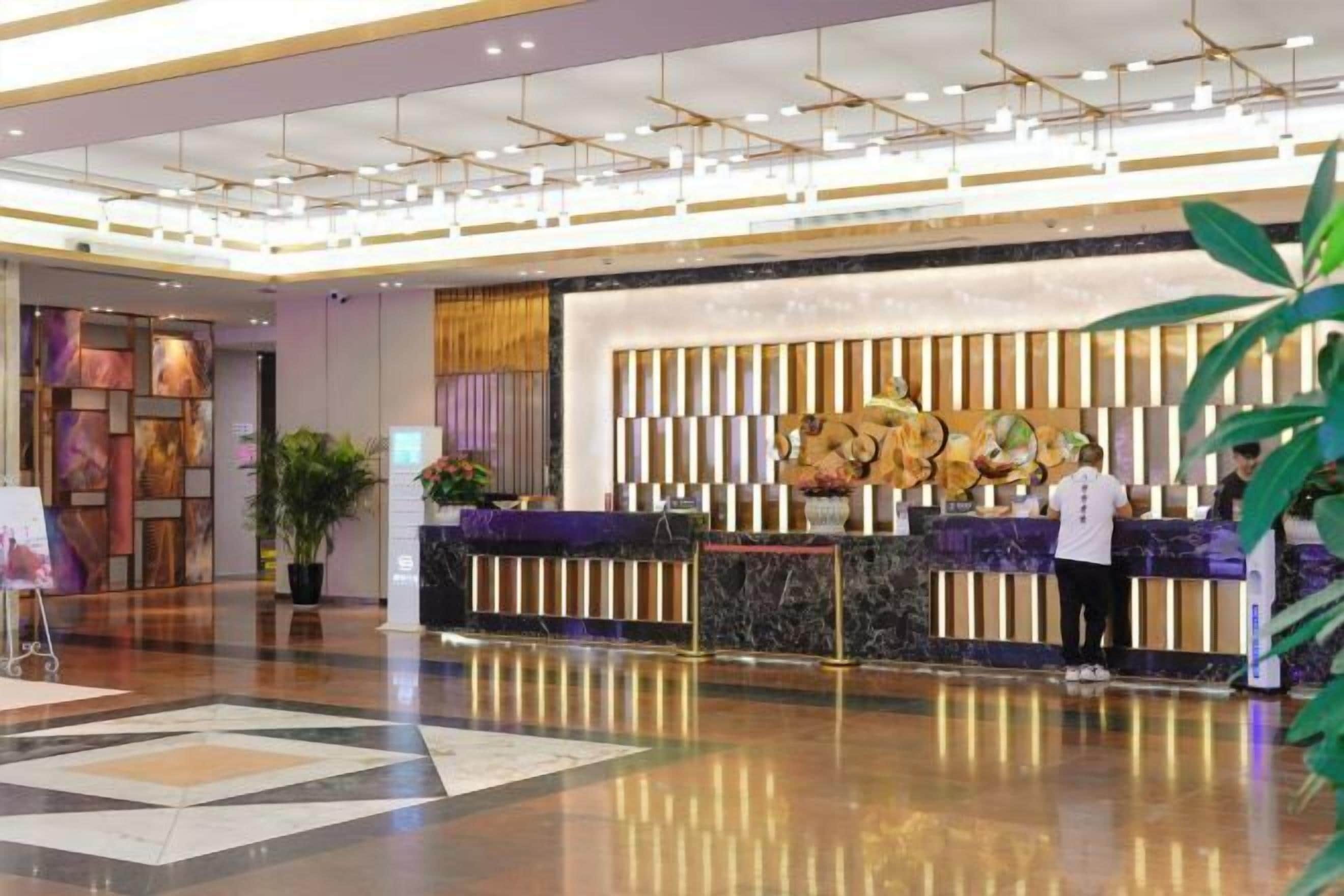 A modern hotel lobby with a reception desk, a large abstract painting, and a potted plant.