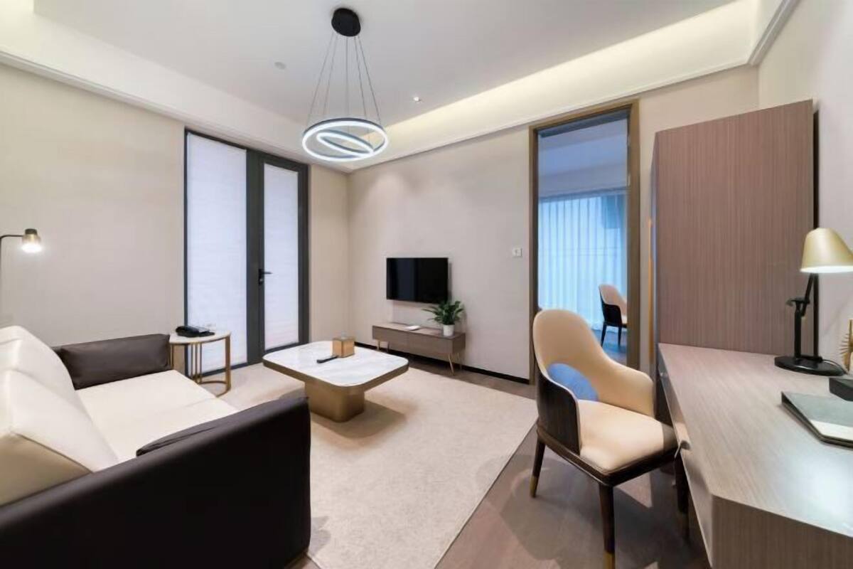 A modern hotel room with a sofa, a desk, a TV, and a large window.