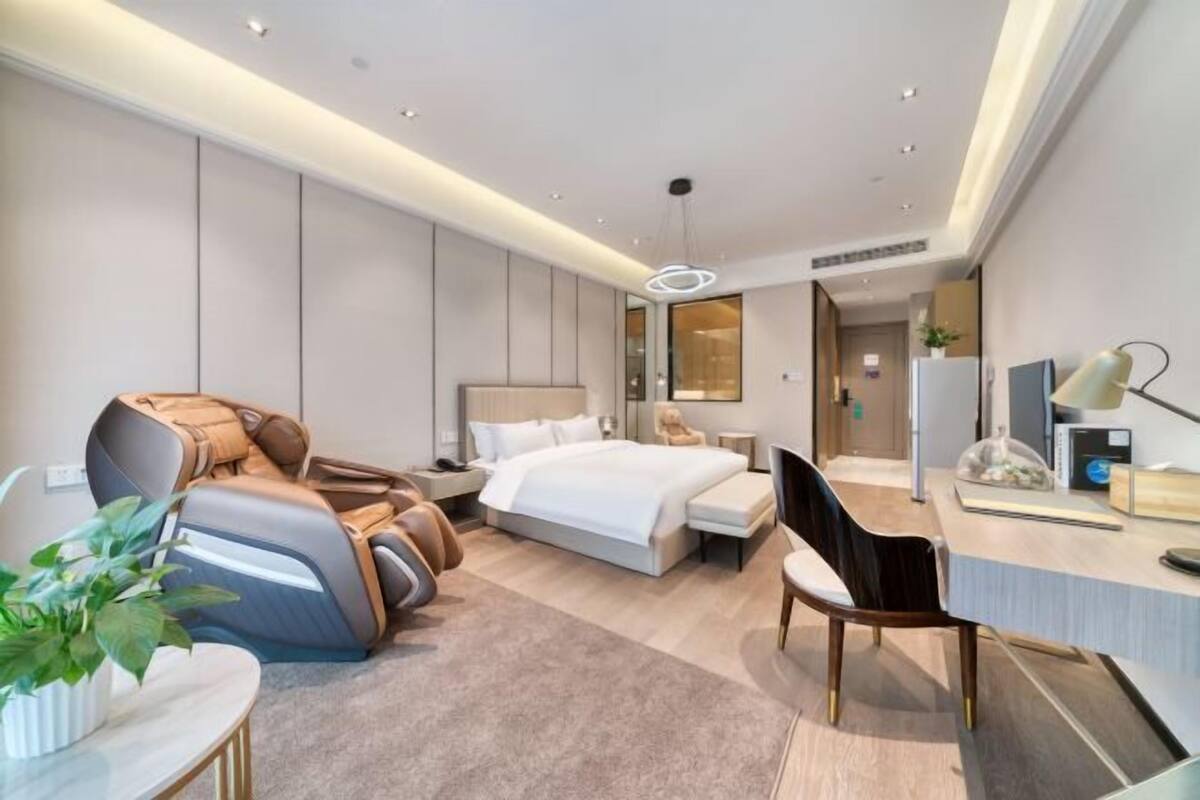 A modern hotel room with a bed, massage chair, desk, chair, and a potted plant.