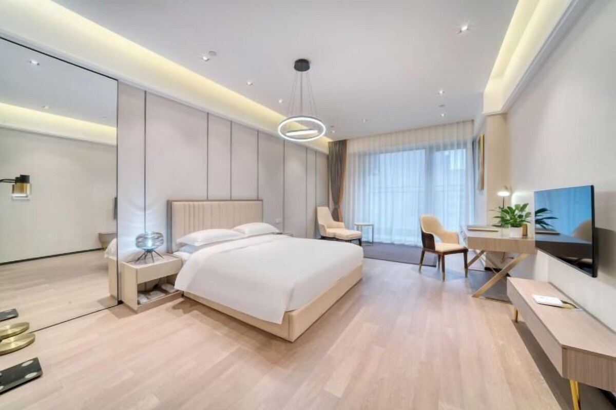 A modern hotel room with a large bed, a desk, a chair, a TV, and a bedside table with a lamp.