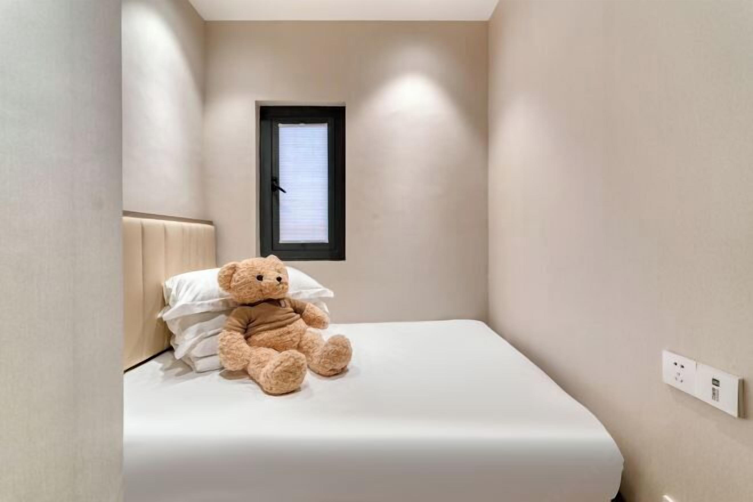 Quarto pequeno com uma cama de solteiro, janela, um urso de pelúcia na cama e uma tomada na parede.