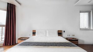 A hotel room with a large bed, a window with blinds, a nightstand, and a view of the city.