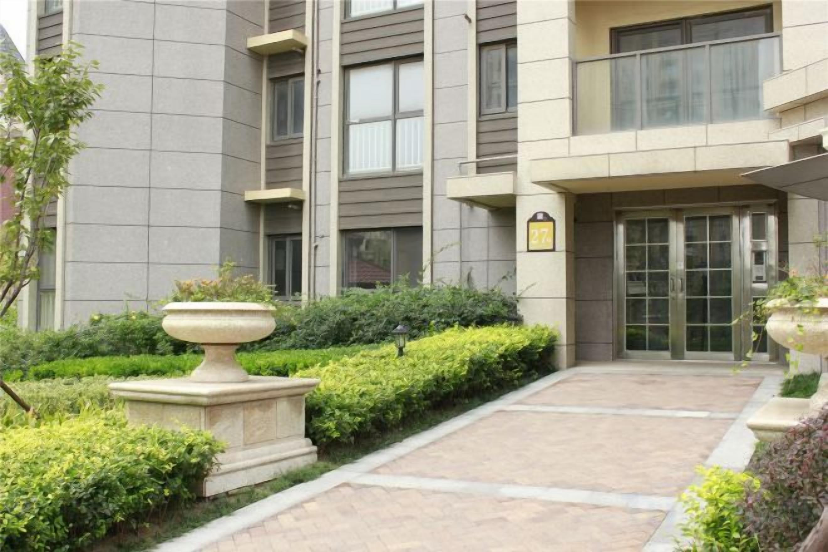 Una entrada de edificio con un camino pavimentado, una jardinera de piedra y una puerta con un letrero en la parte superior.