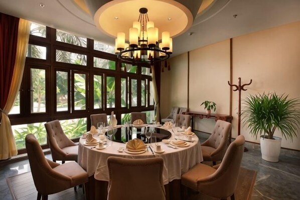 A dining area with a round table set for six, surrounded by chairs, with a large window and a chandelier.