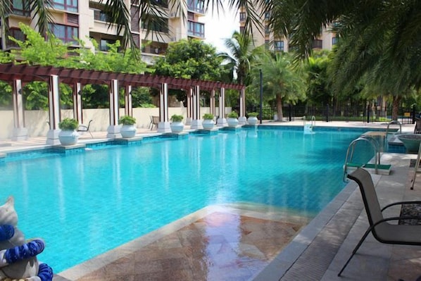 A swimming pool with lounge chairs and a covered seating area, surrounded by greenery and a building in the background.
