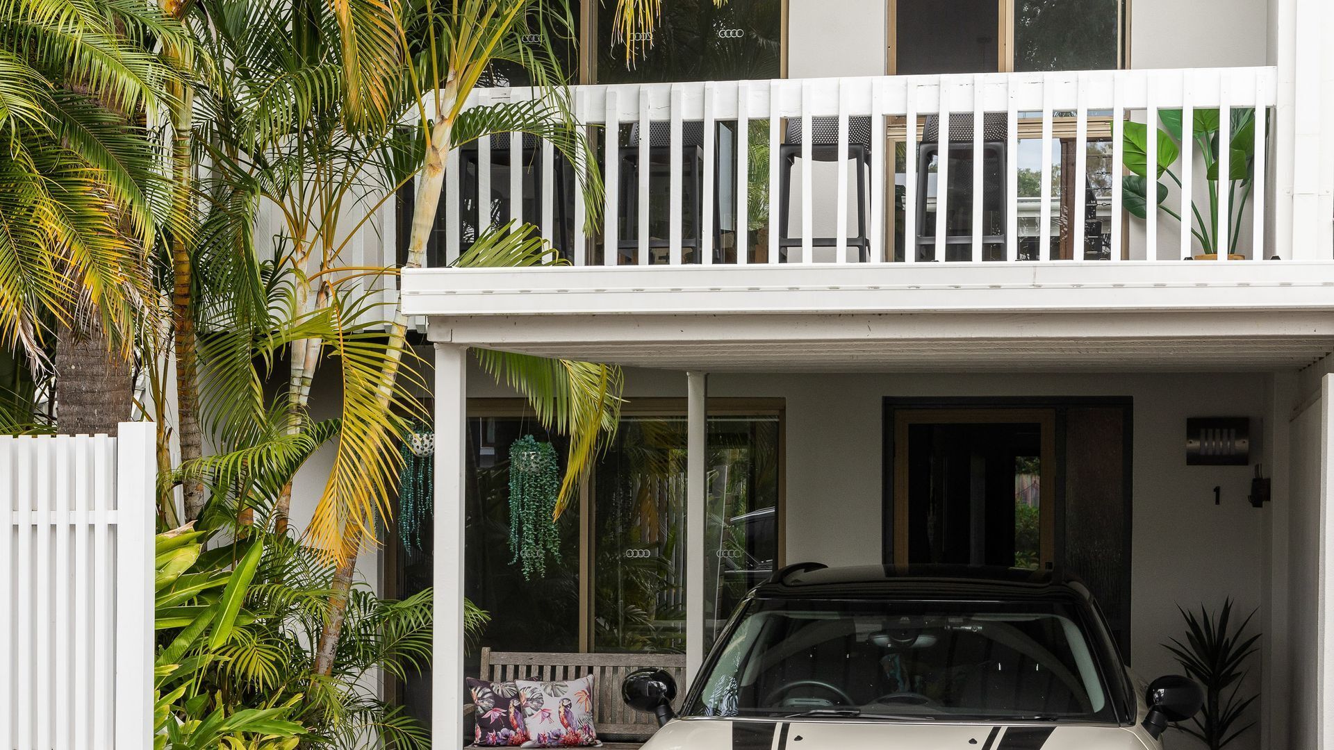 Une maison à deux étages avec un balcon blanc, une voiture garée devant et une végétation luxuriante.