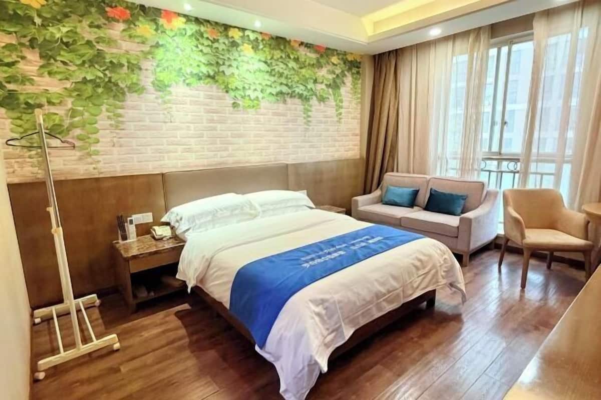 Habitació d'hotel amb llit, sofà, cadira, tauleta de nit, làmpada i paret de maó amb vegetació.
