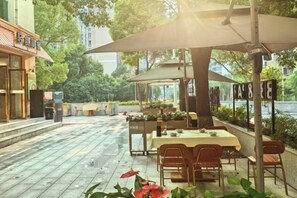 Un área de comedor al aire libre con mesas y sillas, un espacio de estar cubierto y vistas a un edificio y follaje.