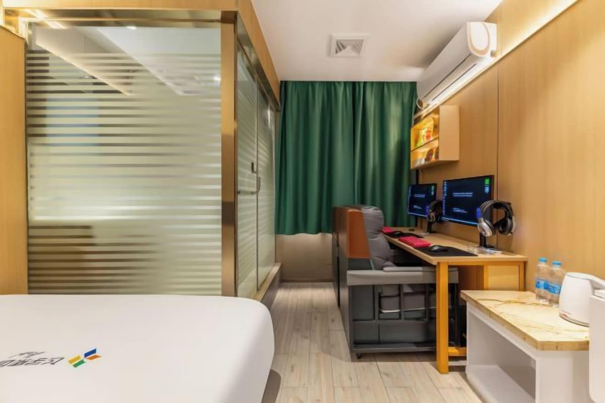 A hotel room with a bed, a desk with a computer, a chair, and a small table with a lamp and bottles.