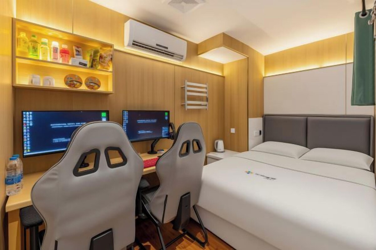 A hotel room with a bed, two desks with computers, a shelf with toiletries, and an air conditioning unit.
