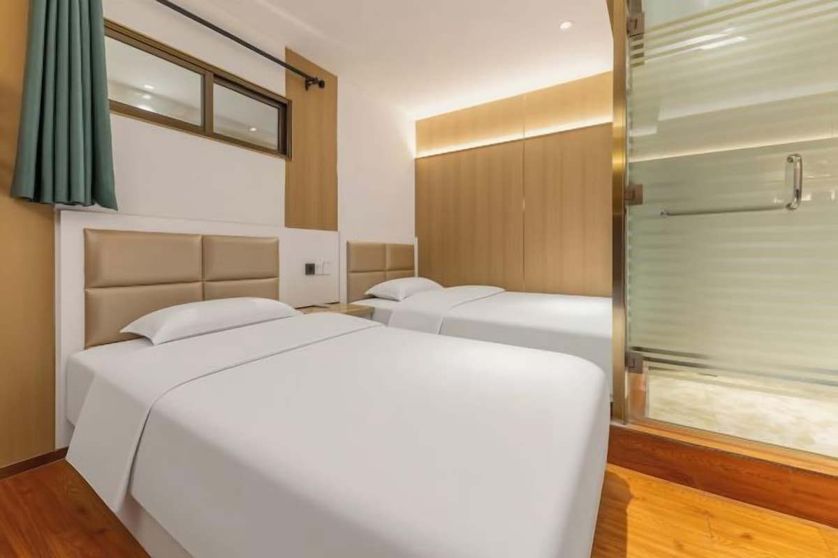 A hotel room with two beds, a large window with curtains, a wooden headboard, and a glass shower enclosure.
