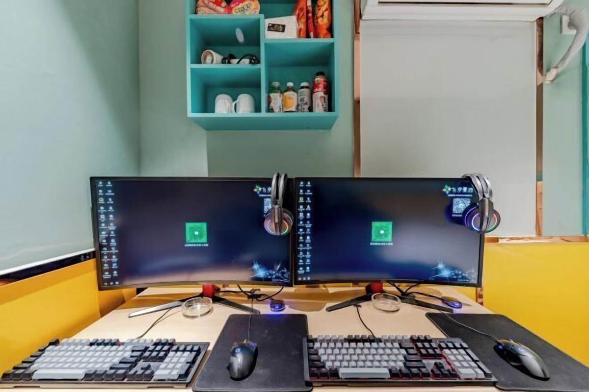 Estació de treball amb dos monitors, dues tecles i dos ratolins, una prestatgeria blau maragda amb diversos objectes i una paret sense decoració al fons.