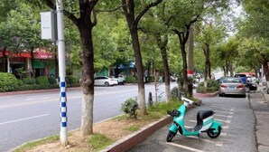 A teal scooter parked on a sidewalk next to a road with trees and buildings in the background. - Hakka Hotel (Huichang)