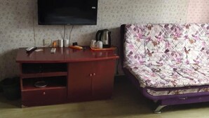 Une pièce avec un téléviseur fixé au mur, un meuble en bois avec des tiroirs, un lit avec une literie à motifs floraux et une bouilloire posée sur le meuble.