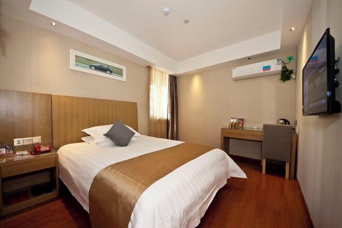 A hotel room with a bed, bedside table, desk, chair, TV, and a wall-mounted shelf.