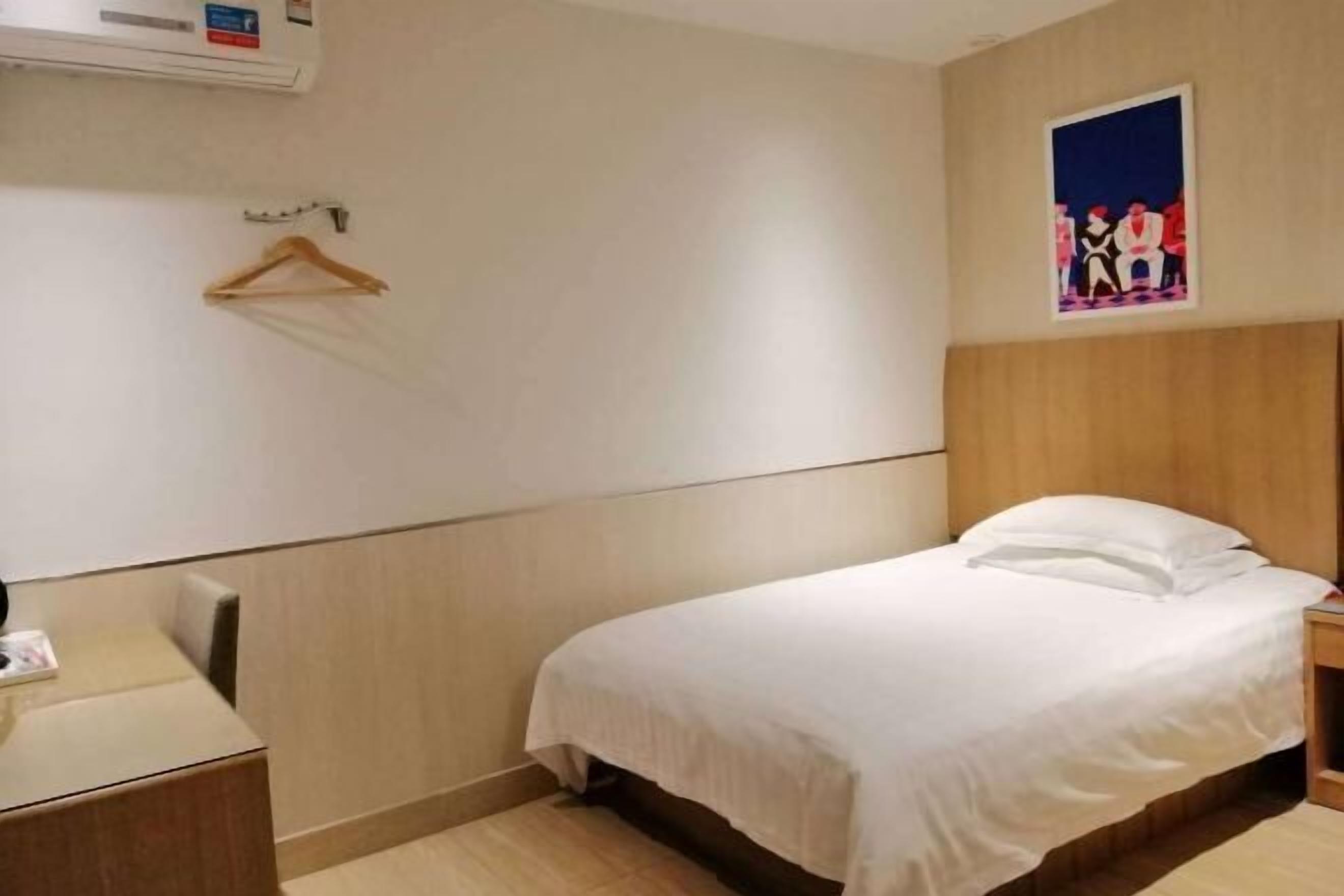 A hotel room with a bed, a desk, a chair, a coat rack, and a painting on the wall.
