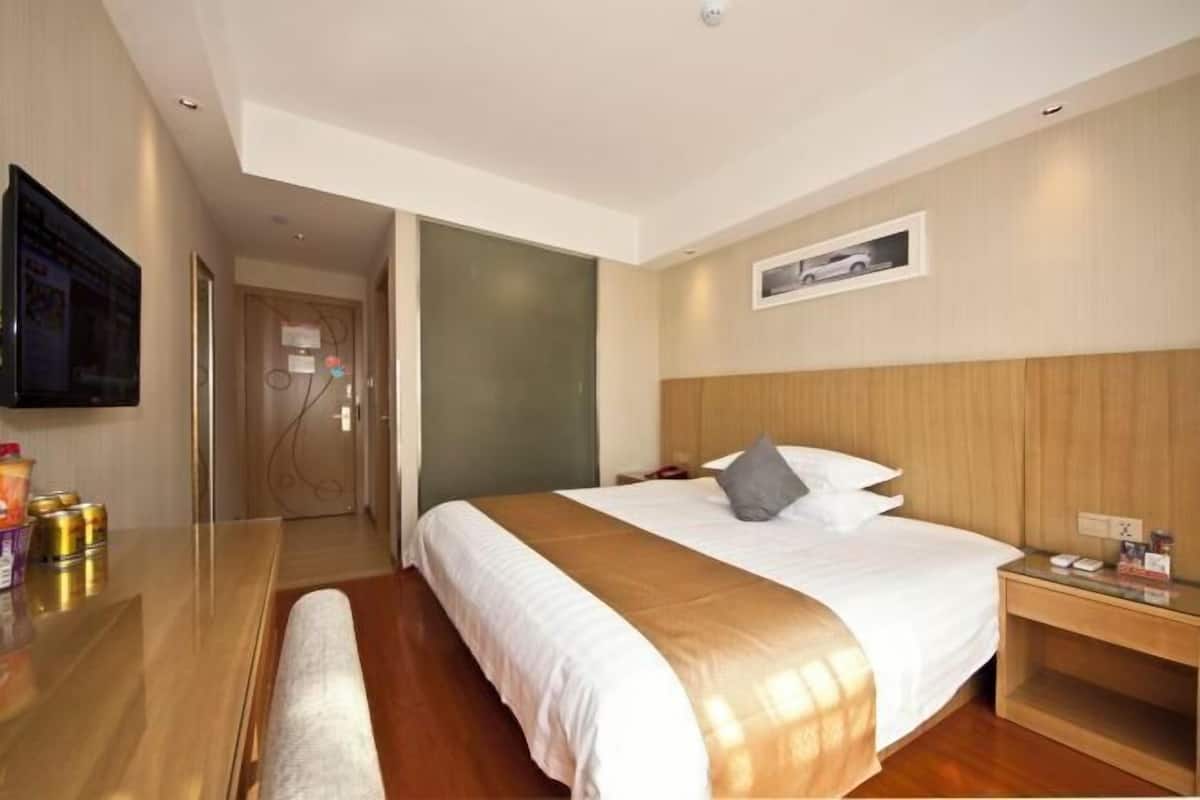 A hotel room with a large bed, wooden headboard, a bedside table, a flat-screen TV mounted on the wall, and a door leading to another room.
