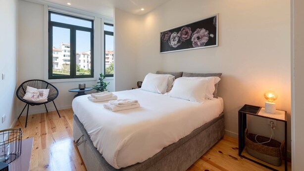 Een moderne slaapkamer met een groot bed, een stoel, een nachtkastje met een lamp en een raam met uitzicht op de stad.