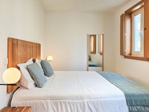 Un dormitorio con cabecera de madera, cama con ropa blanca, mesita de noche con lámpara y ventana con vistas al exterior.