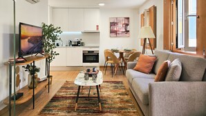 Ein modernes Wohnzimmer mit grauem Sofa, Couchtisch und Fernseher auf einem Schrank. Eine Küchenzeile mit weißen Schränken und ein Esstisch für vier Personen.