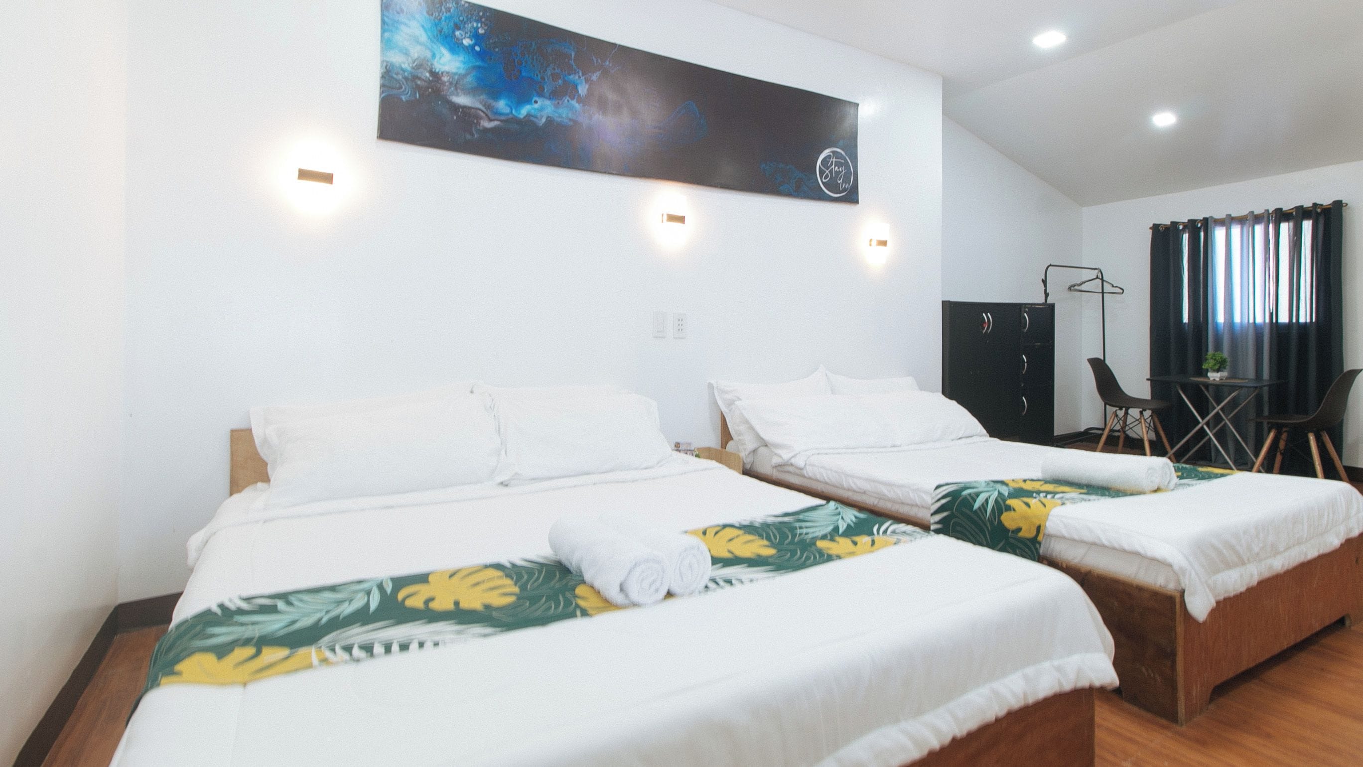 A hotel room with two beds, a painting on the wall, a desk with a chair, and a wardrobe.