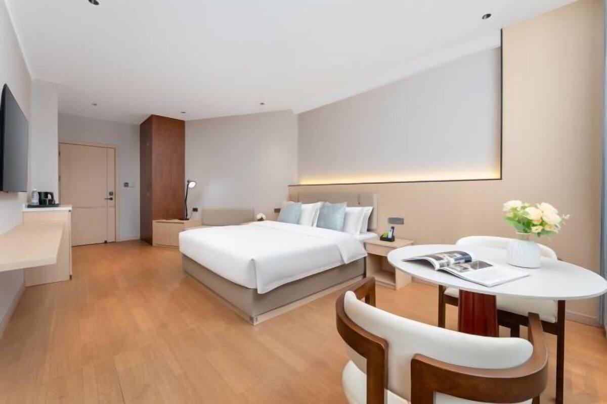 Moderni hotellihuone, jossa on suuri sänky, yöpöydät, työpöytä tuolilla sekä pieni pyöreä pöytä, jonka päällä on maljakko täynnä kukkia.