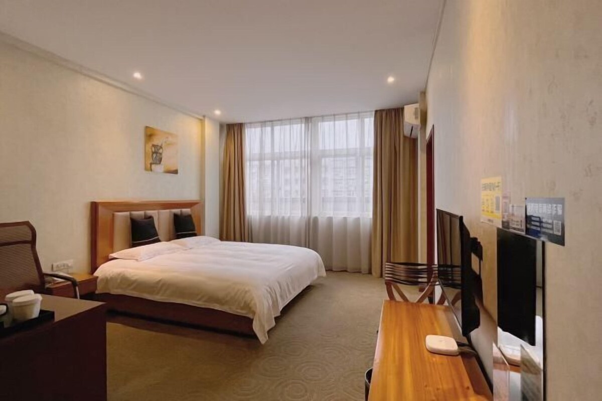 Ein Hotelzimmer mit Bett, Schreibtisch, Stuhl, Fernseher und einem fensterbelaubten Fenster.
