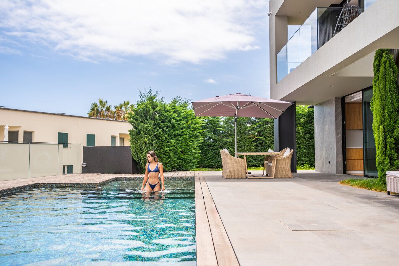 Uma área moderna de piscina ao ar livre, com uma pessoa de biquíni, uma mesa com cadeiras e um guarda-sol.