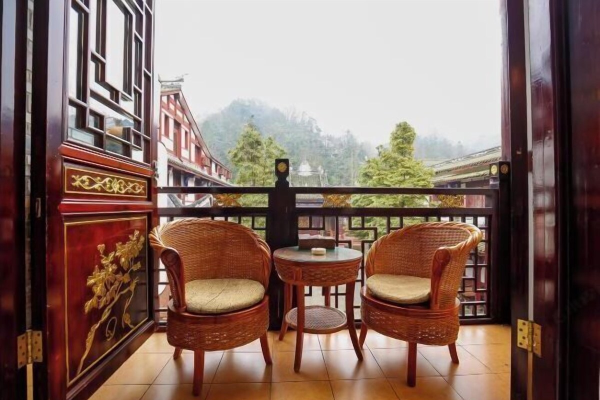 Ein Balkon mit Korbstühlen und einem kleinen Tisch, mit Blick auf ein traditionelles Gebäude und Bäume.