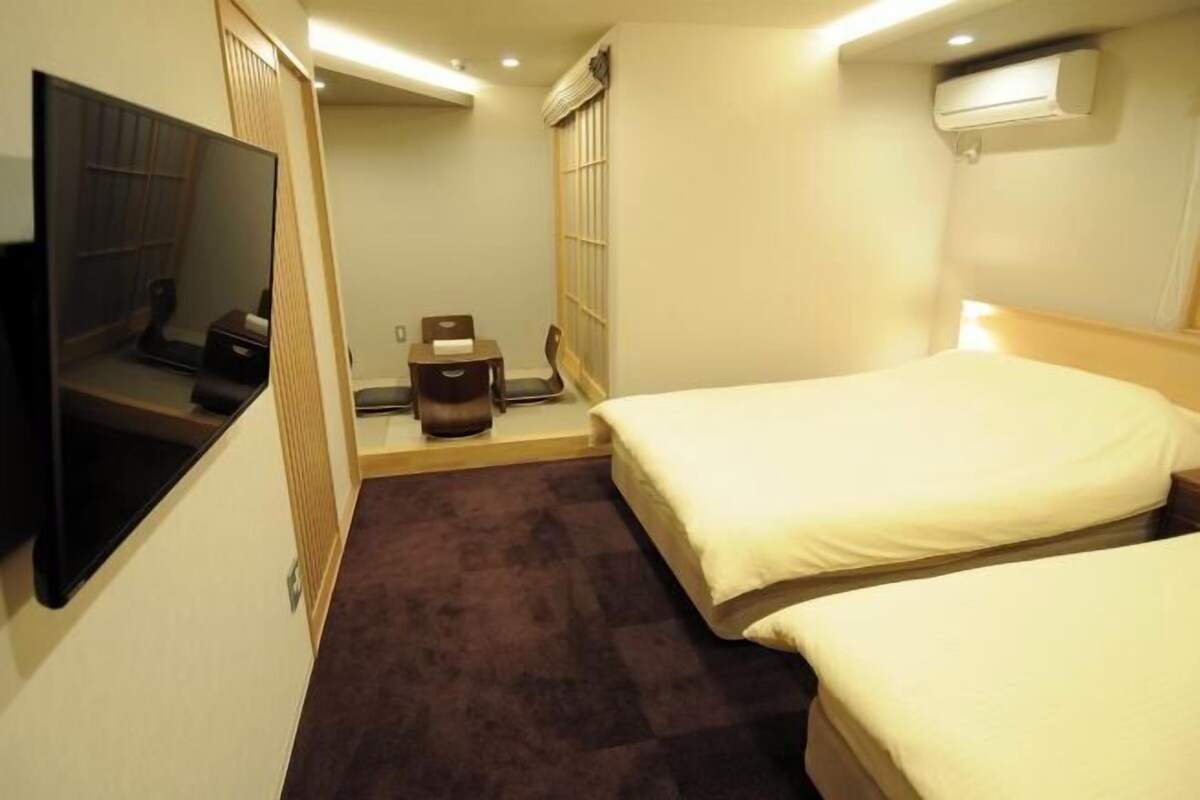 A hotel room with two beds, a TV, a small table, and a window with blinds.