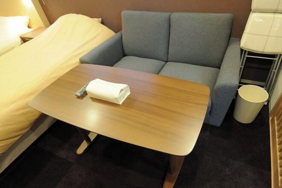 A compact hotel room with a bed, a small sofa, a wooden table, and a trash bin.