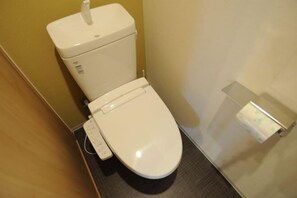 A white toilet with a flush handle and a bidet attachment in a small bathroom with a plain wall and a wooden floor. - Hiz Hotel Kyoto-Nijo Castle (Kyoto)
