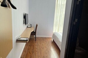 A narrow hallway with a wooden floor, a bed with white bedding, a chair, a small table with a glass container, and a mounted television. - Hexingshun Hotel (Chengdu)