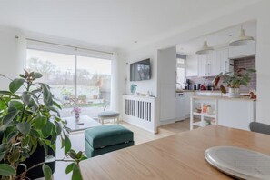 A modern kitchen with white cabinets, a dining area with a wooden table, and a large window overlooking a garden.
