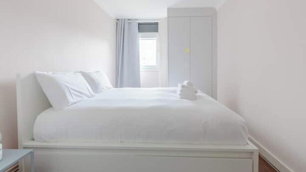 Una cama blanca en una habitación con ventilador de techo, una ventana con cortinas y un armario blanco.