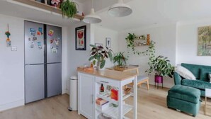 Una cocina moderna con un refrigerador empotrado, una mesa de madera y un sofá verde en un salón luminoso con plantas y obras de arte.