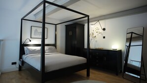 Een hemelbed met donker frame, een nachtkastje met lamp, een klein bureau met spiegel, en een lijst met een afbeelding aan de muur.