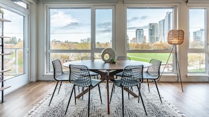 A dining area with a round table, six chairs, and a view of a cityscape through large windows. - Sophari Parkside: Bellevue Apartments (Bellevue)