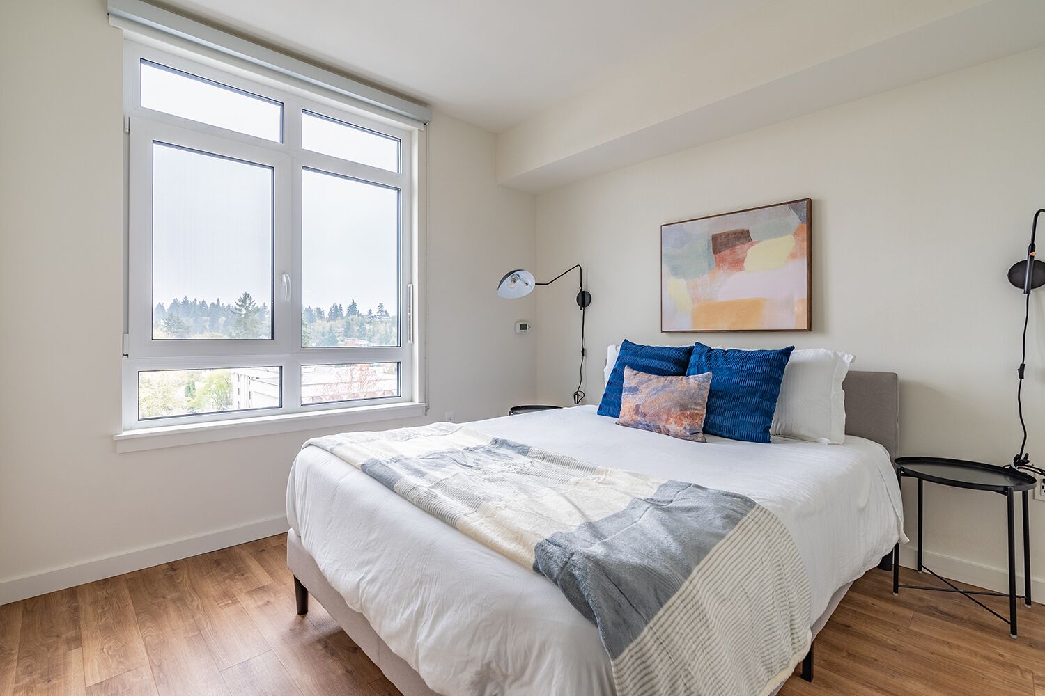 Une chambre avec une grande fenêtre, un lit recouvert d’une courtepointe, deux tables de chevet et une lampe fixée au mur.